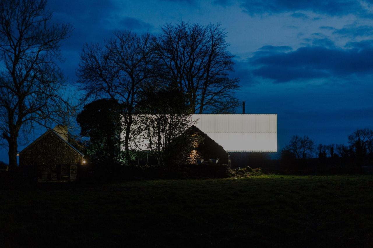 Exterior noapte cu container alb luminat si casa veche din piatra in fundal