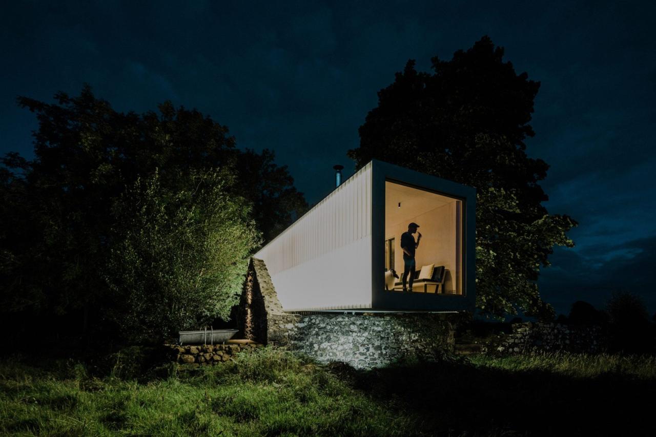 Exterior noapte cu container alb luminat, persoana vizibila in fereastra panoramica, pe fundatia de piatra