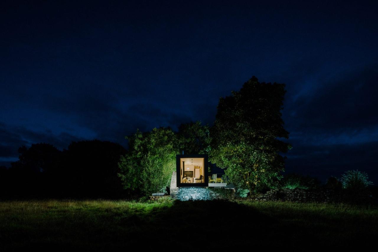 Exterior noapte cu container negru luminat din interior, persoana vizibila la masa, pe fundatia de piatra iluminata