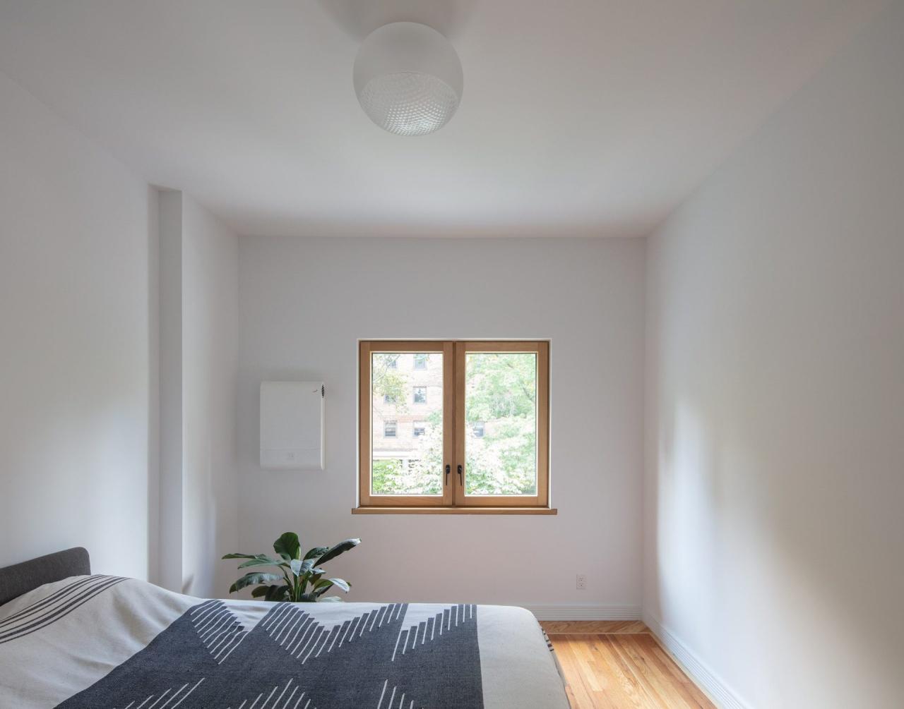 Dormitor minimalist alb cu pat gri, fereastra dubla cu rama din lemn si lampa sferica alba la tavan - unitate de ventilatie pe perete