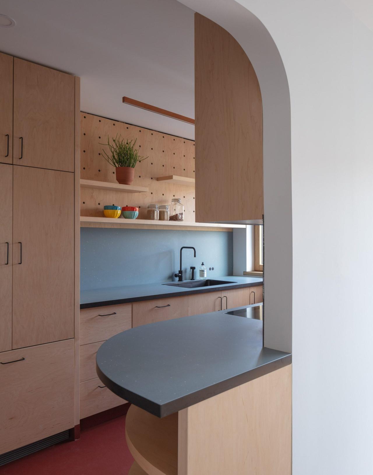 Bucatarie cu dulapuri din lemn deschis, blat gri albastru, backsplash albastru, rafturi cu pegboard si arc rotunjit spre intrare