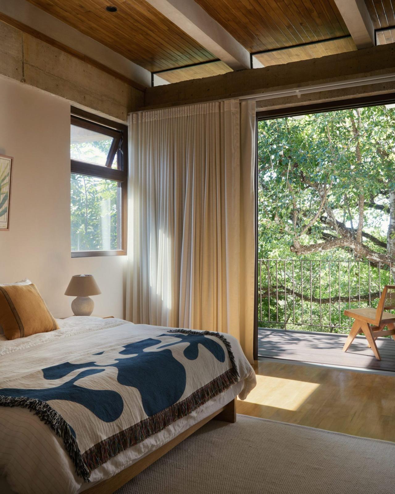 Dormitor cu pat simplu, cuvertura albastra cu franjuri, usa glisanta spre terasa cu vedere la copaci – foto Thibault Cartier / Dezeen