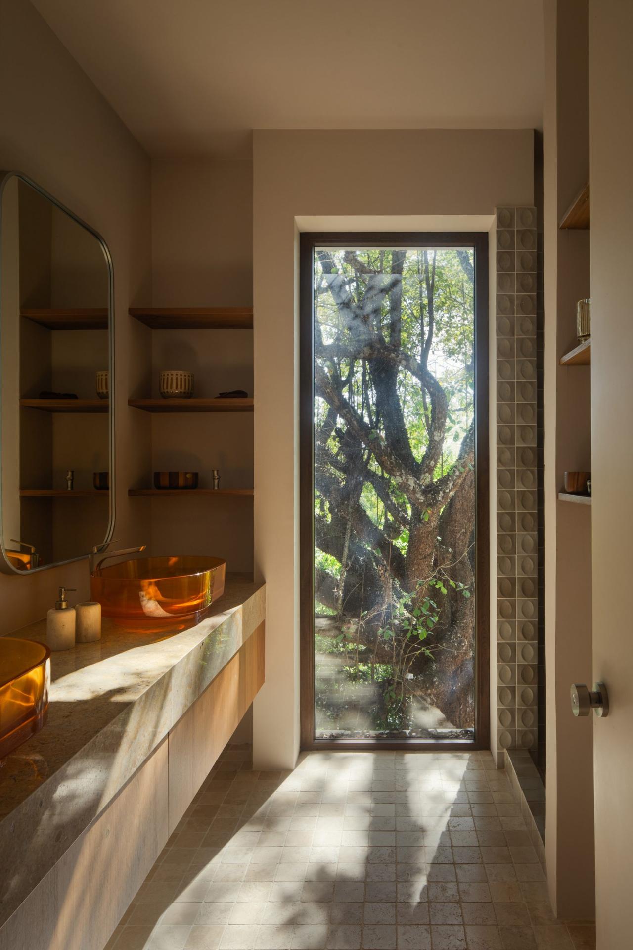 Baie cu chiuveta din sticla portocalie pe blat de piatra, fereastra inalta cu vedere directa la copacul pochote – foto Thibault Cartier / Dezeen