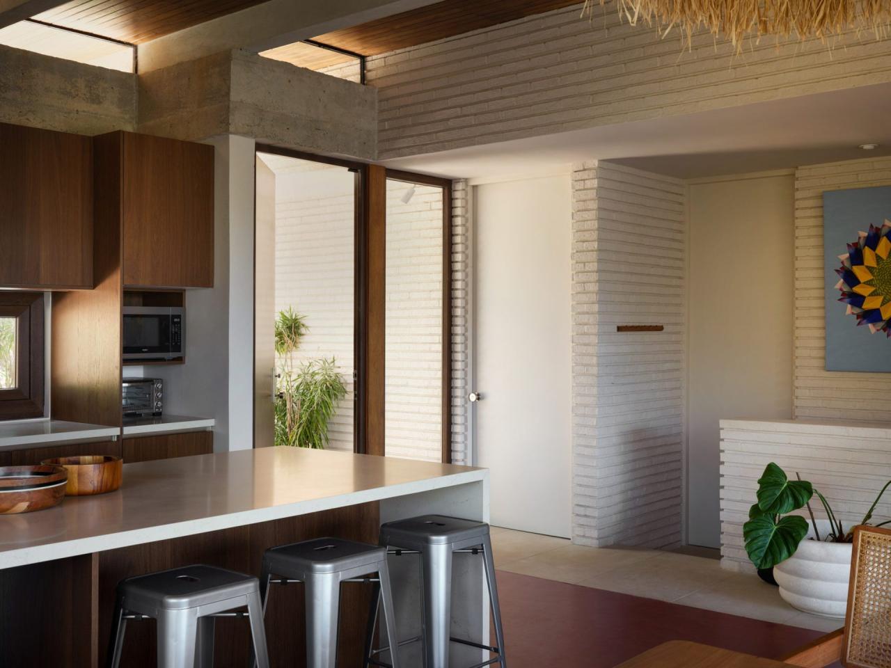 Bucatarie open-space cu insula alba, dulapuri din lemn inchis si vedere spre curtea interioara cu plante – foto Thibault Cartier / Dezeen