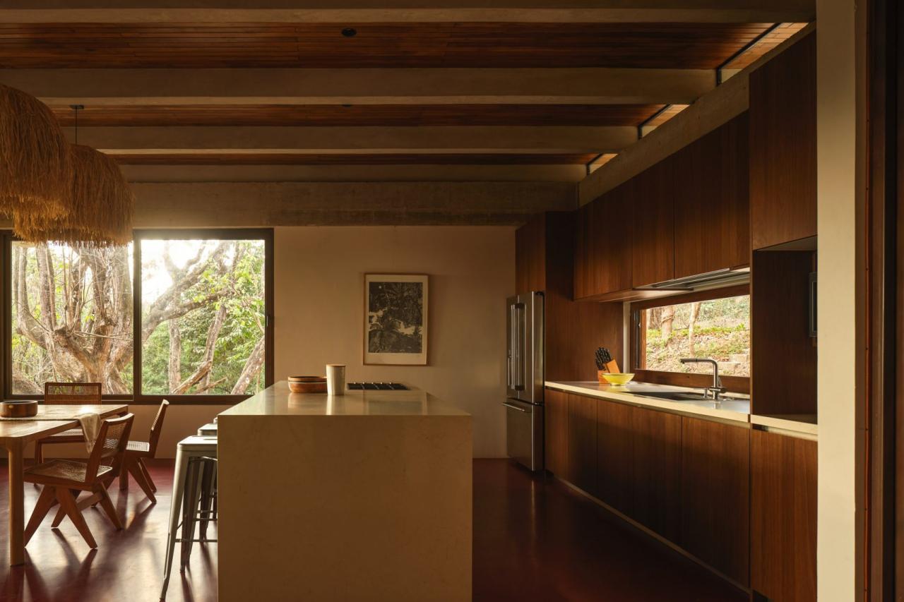 Bucatarie cu dulapuri din lemn inchis, insula cu blat alb si fereastra panoramica cu vedere la copaci – foto Thibault Cartier / Dezeen