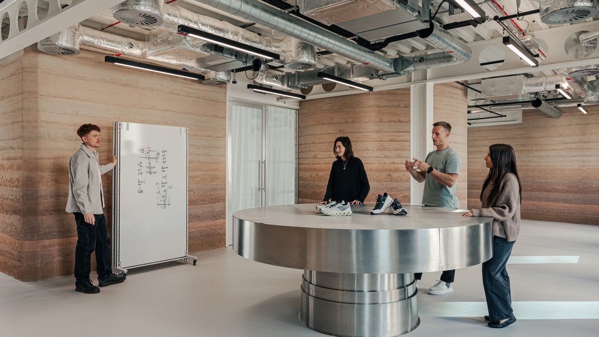 Zona de colaborare cu masa rotunda din inox si perete din pamant compactat, echipa in discutie langa whiteboard mobil – foto Chris Wharton / M Moser Associates