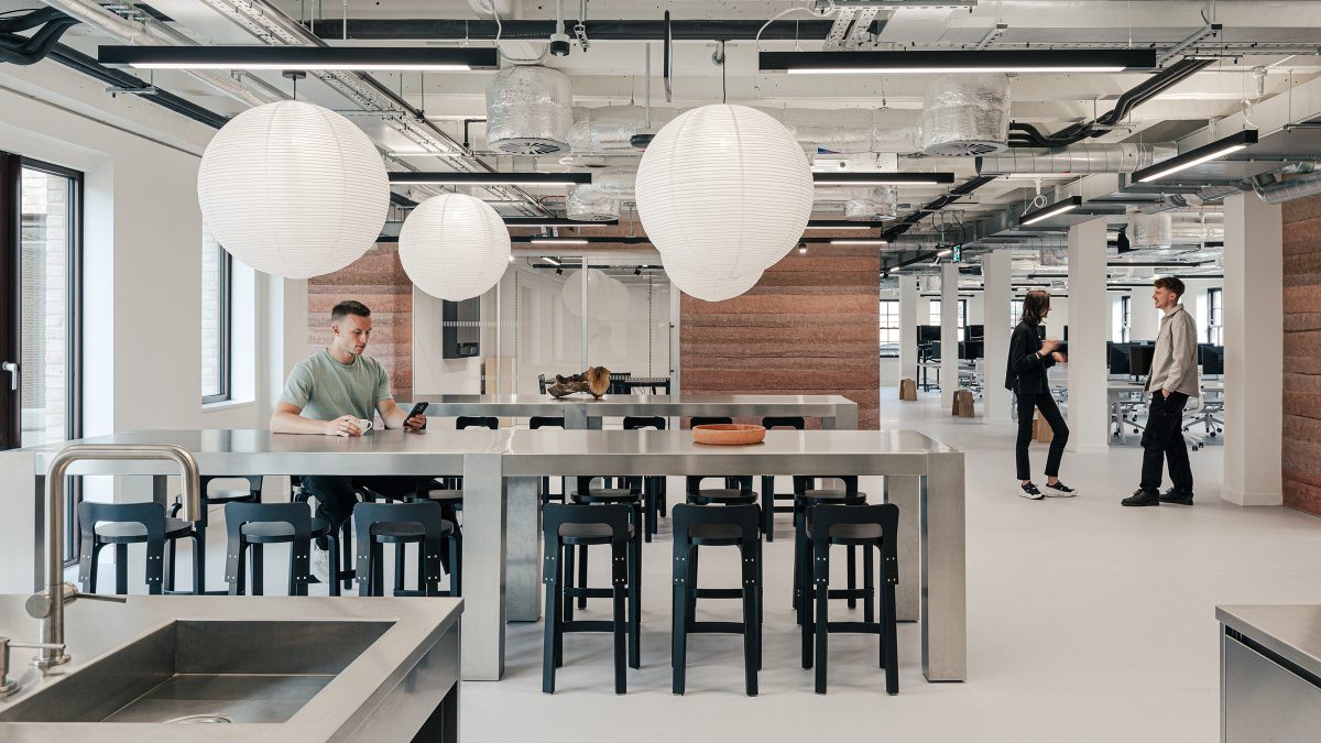 Zona de dining/tea point cu mese din inox, scaune bar negre si lampe suspendate albe, cu zona de lucru in fundal – foto Chris Wharton / M Moser Associates
