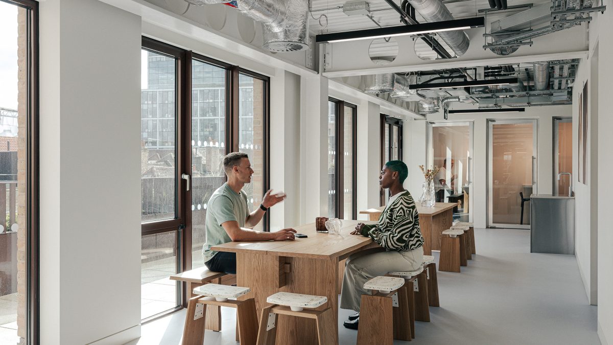 Zona de lucru informal cu mese si scaune din lemn de stejar, ferestre mari si culoare cu birouri private in fundal – foto Chris Wharton / M Moser Associates