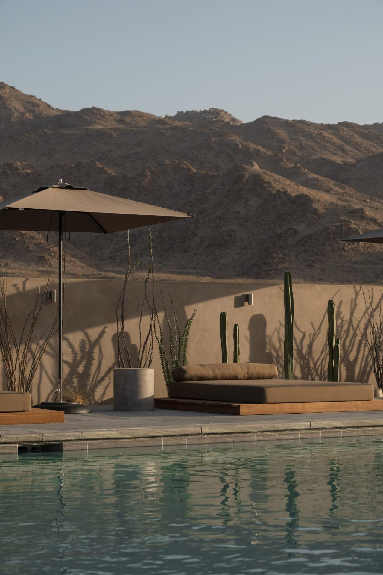 Piscina cu sezlonguri pe platforme din lemn, cactusi si perete de lut in lumina apusului